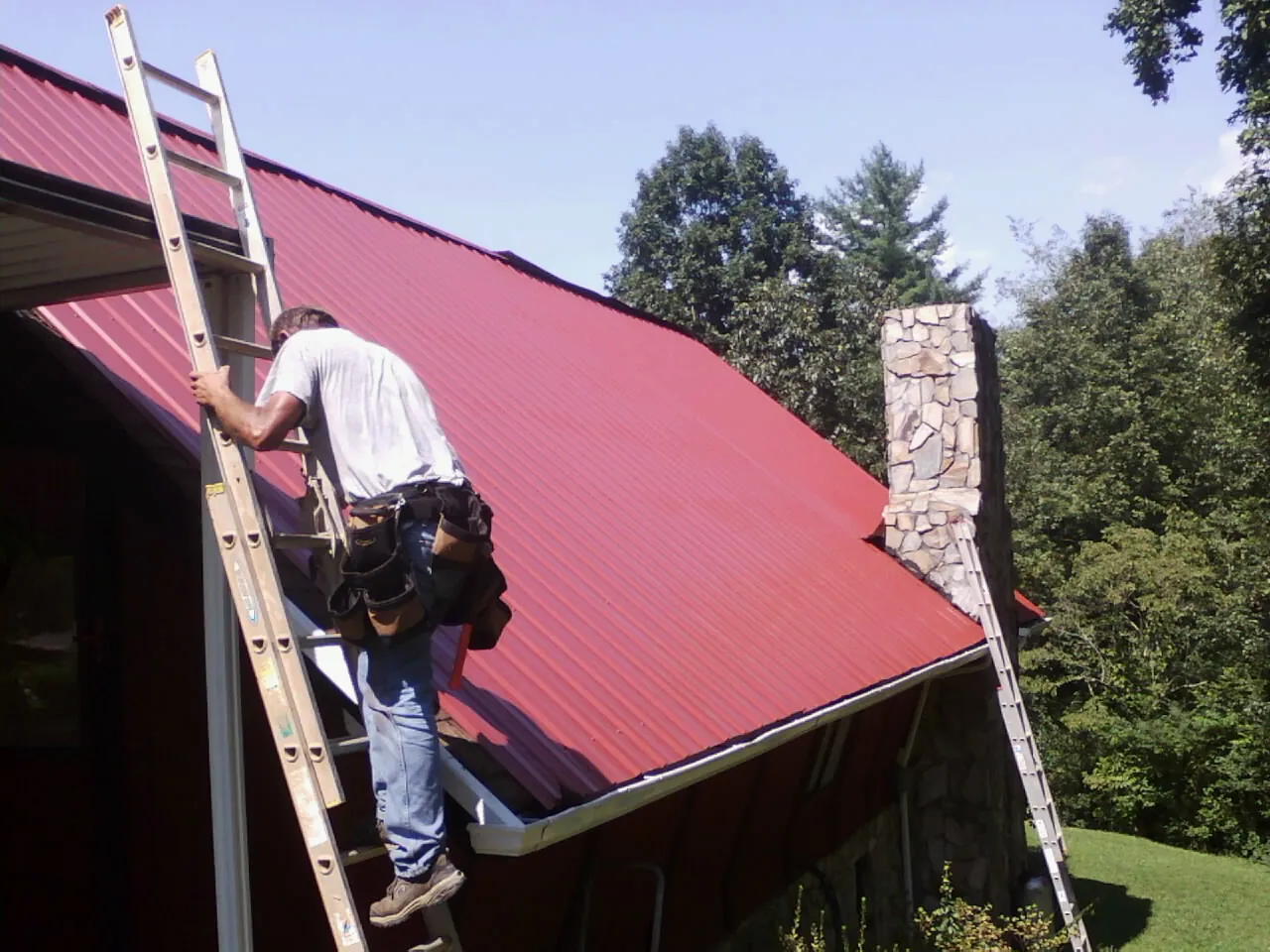 Roofer on a ladder beside a red metal roof and stone chimney — Soffit & Fascia Repair by Roofing Brothers in Ruston